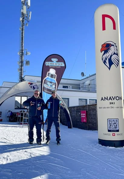 Two men in matching Anavon Ski jackets standing in the snow at a resort, holding water bottles in front of a Powerade banner and an Anavon branded inflatable pillar with a QR code.